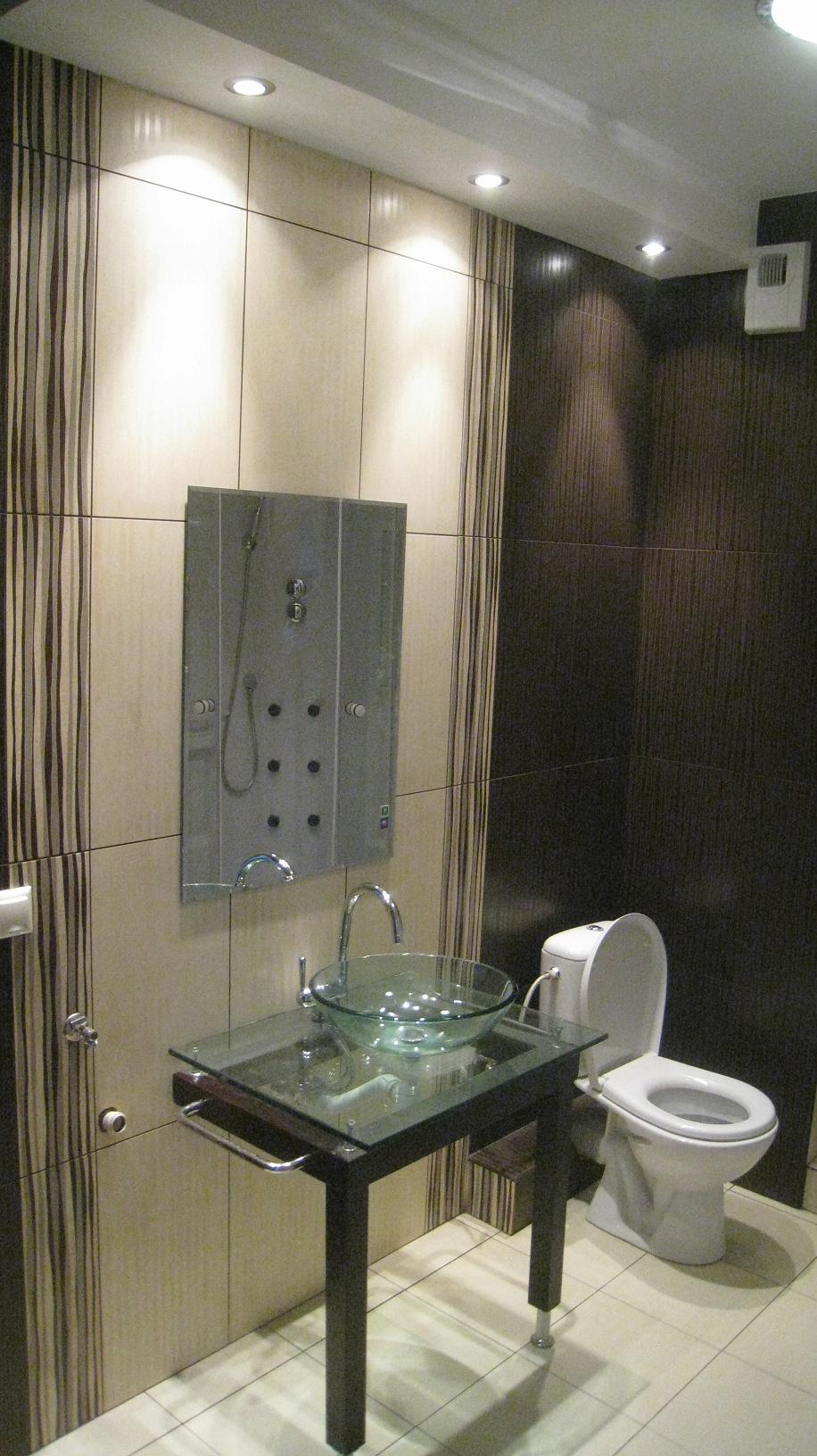 Nowoczesna łazienka z kabiną prysznicową z hydromasażem, umywalką z przezroczystego szkła na czarnym stoliku z metalowymi nogami, toaleta w tle i płytki w odcieniach beżu i brązu z pionowymi paskami.